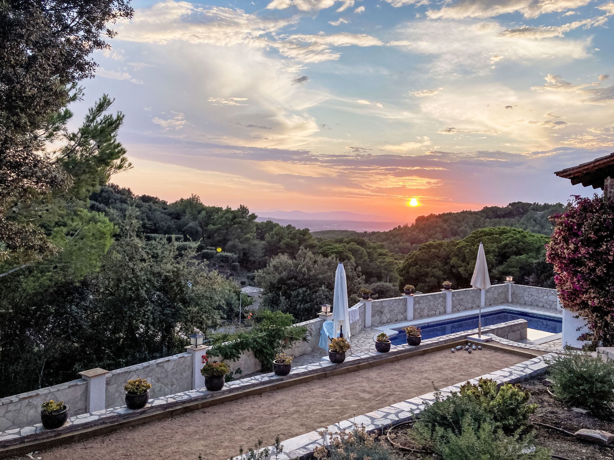 A pool area with a stone wall and potted plants, surrounded by trees and a sunset view.