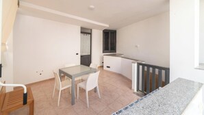 Un pequeño salón de planta abierta con mesa y sillas, zona de cocina con mostrador y ventana, y escalera con barandilla.