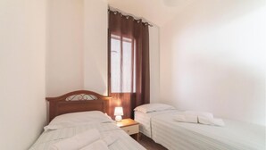 Una habitación pequeña y limpia con dos camas individuales, una mesita de noche con lámpara y una ventana con cortinas.