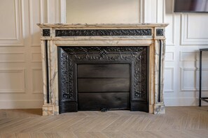 Une cheminée ornée de sculptures complexes, flanquée de manteaux de cheminée en marbre, dans une pièce aux murs lambrissés et au parquet disposé en motif chevrons.