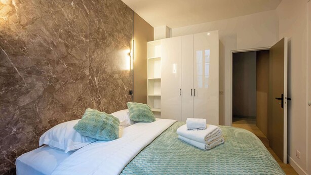 Ein modernes Schlafzimmer mit Marmorwand, einem Bett mit weißen Leinentextilien, grünen Kissen und einer blauen Decke. Weiße Schränke und eine Tür, die in einen anderen Raum führt, sind ebenfalls zu sehen.
