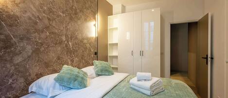 Une chambre moderne avec un mur en marbre, un lit recouvert de draps blancs, des coussins verts et une couverture bleue. On y trouve des armoires blanches et une porte visible qui mène à une autre pièce.