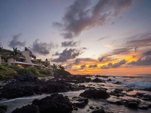 Una località balneare con costa rocciosa, palme e vista sul tramonto.