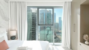 A modern hotel room with a large bed, a TV, and a view of high-rise buildings through floor-to-ceiling windows. - Burj Royale Elegance Cityscape From Your Balcony (Dubai)
