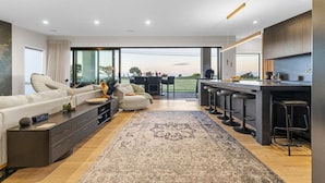 Una sala moderna con un sofá, una mesa de centro y una alfombra. Cuenta con una zona de cocina con una barra y taburetes. La habitación tiene ventanas grandes y vistas al exterior.