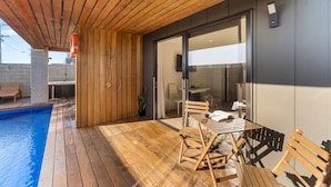 Una terraza moderna al aire libre con piscina, muebles de madera y una puerta de vidrio que da a un espacio interior.