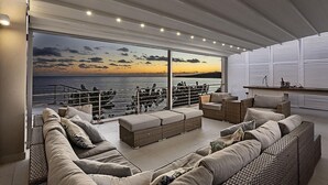 A spacious balcony with a view of the sea at sunset, furnished with a sofa, ottoman, and wicker chairs.