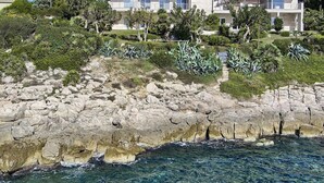 A coastal property with modern architecture, featuring multiple levels and large windows, surrounded by lush greenery and rocky terrain.