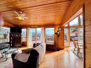 Uma cabana acolhedora com interior de madeira, um ventilador de teto, uma televisão, uma mesa de vidro, um sofá e janelas amplas com vista para o exterior.