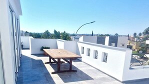 Una terraza en la azotea con una mesa y sillas de madera, paredes blancas y una vista despejada del horizonte de la ciudad.
