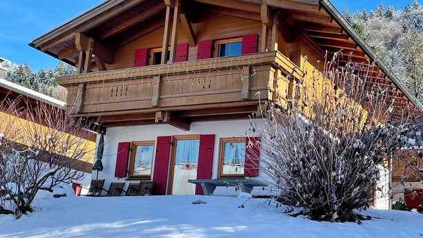 A chalet with a wooden exterior, a balcony, and a stone wall in front.