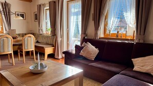 A cozy living room with a sofa, chairs, a wooden table, and large windows with curtains.