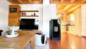 A spacious living area with a wooden ceiling, a television, a refrigerator, and a kitchen in the background.