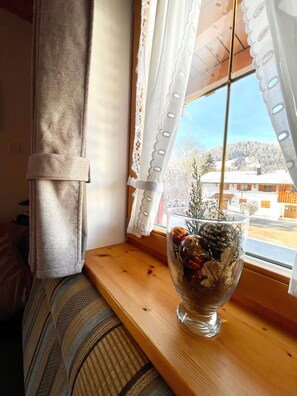 A window with a view of a snowy landscape, a clear sky, and a wooden shelf with a glass vase containing decorative items.
