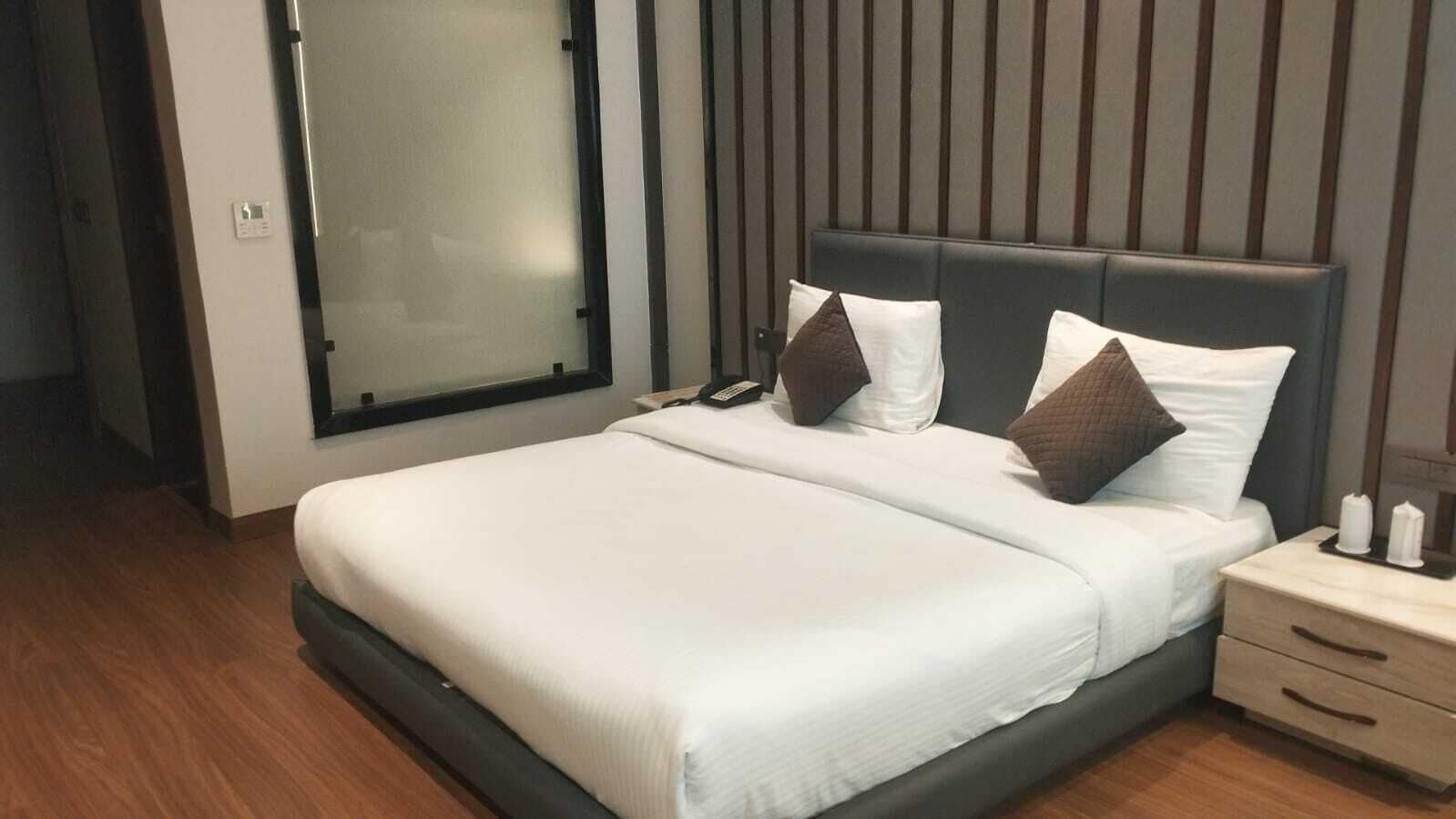 Ein modernes Hotelzimmer mit einem großen Bett, einem Nachttisch mit Telefon und Kerzen sowie einem Fenster mit Jalousien.