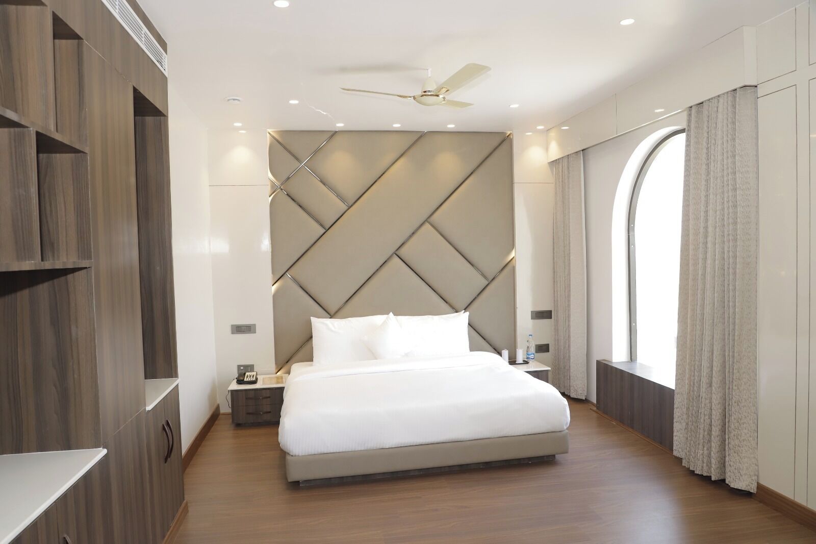 A modern hotel room with a large bed, a wooden wardrobe, a nightstand, and a ceiling fan.