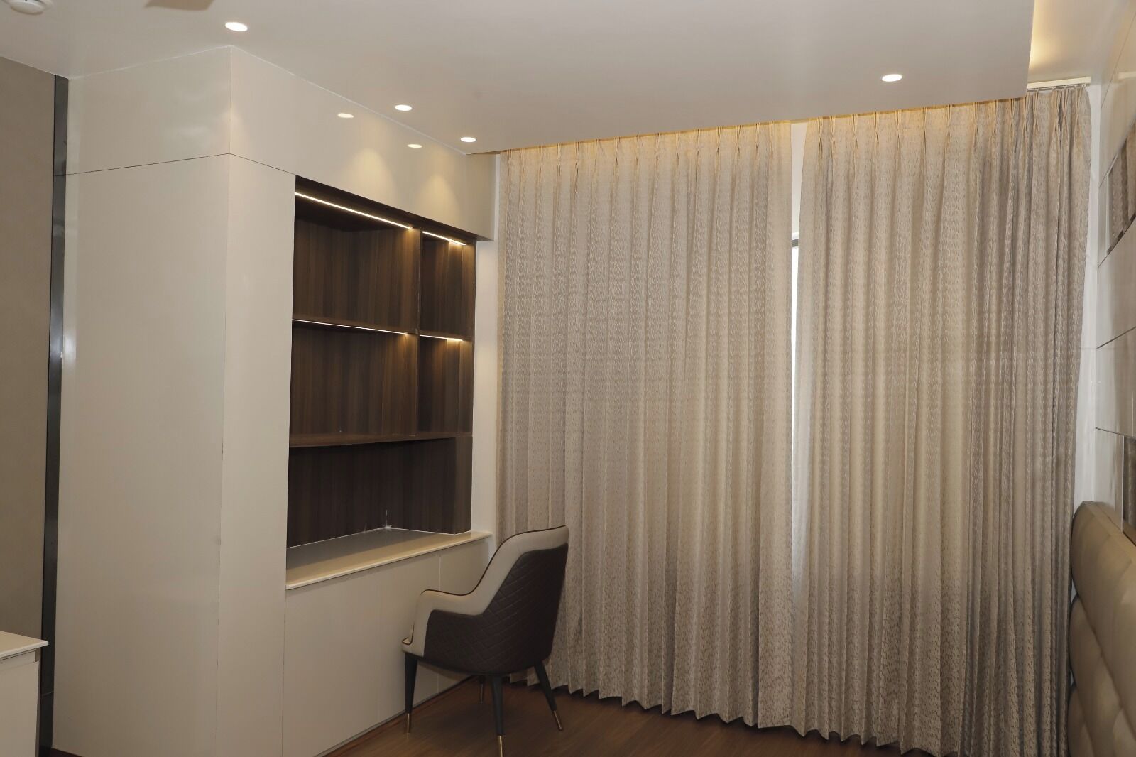 A modern room with built-in wooden shelves, a large window with curtains, and a cushioned chair.