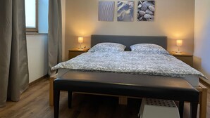 A bedroom with a bed, two bedside tables with lamps, a bench, a window with curtains, and three wall-mounted artworks.