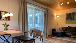 A cozy living room with a wooden ceiling, a dining table with chairs, a sofa, and a painting on the wall.