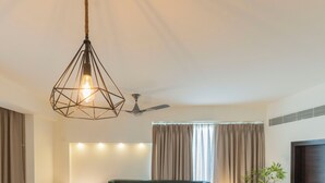 Habitación de hotel con una cama tapizada de verde, dos lámparas de noche, una lámpara colgante geométrica, un ventilador de techo y una ventana con cortinas.