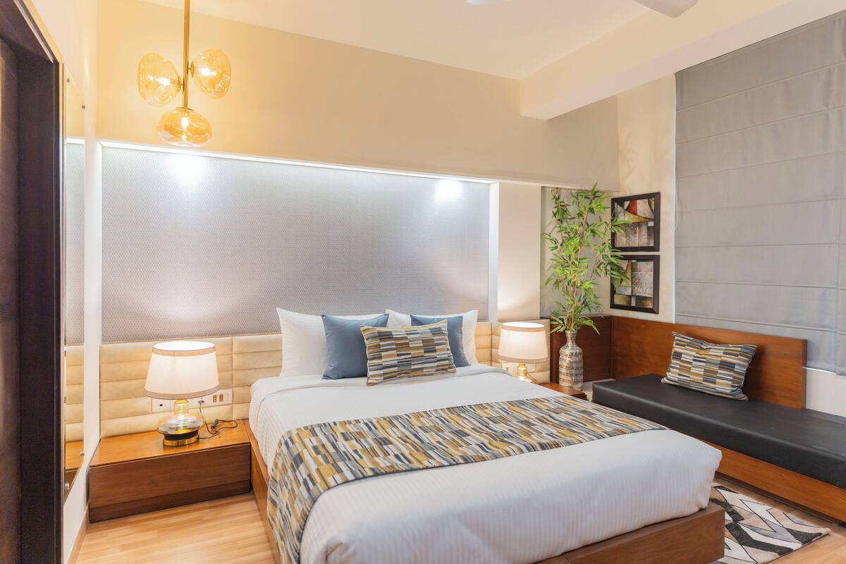 A modern bedroom with a large bed, bedside lamps, a bedside table, a small sofa, and a large window with blinds.