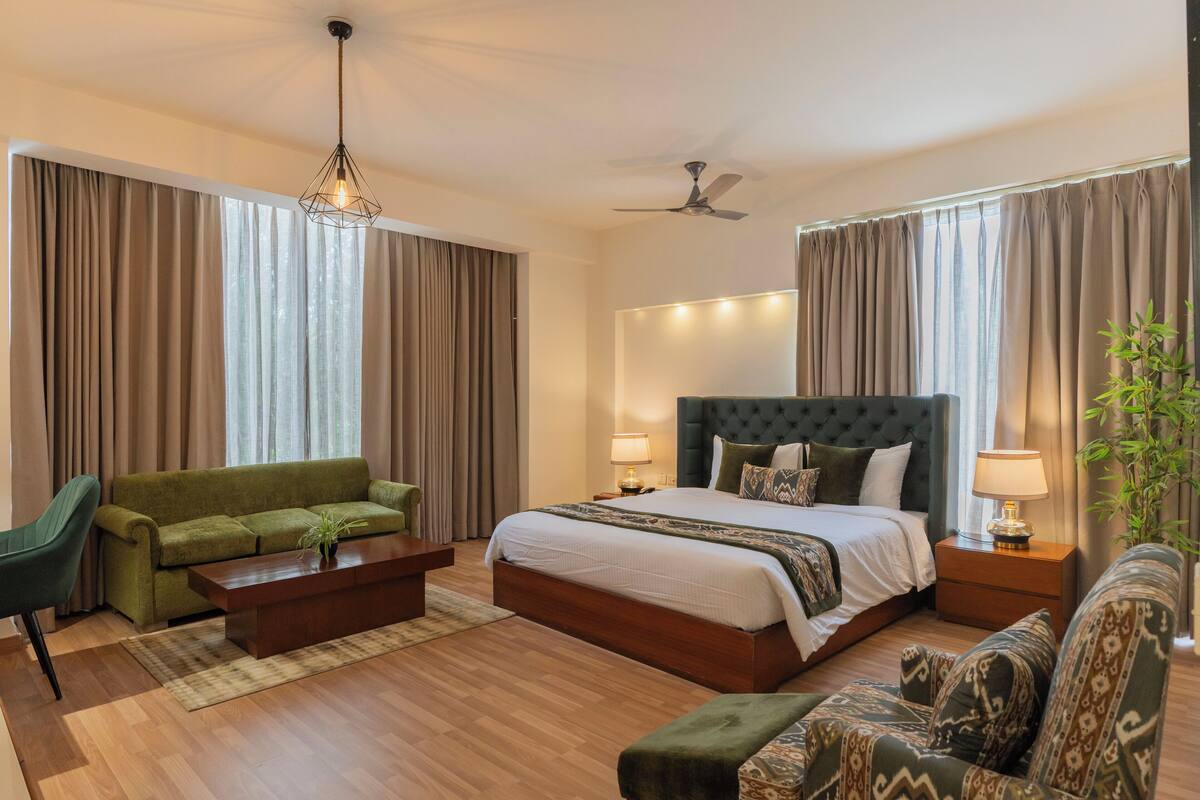 A modern hotel room with a large bed, a green sofa, a wooden coffee table, and a ceiling fan.