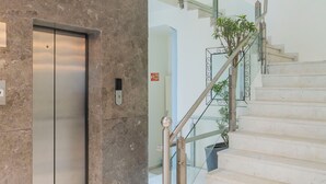 Un ascensor moderno, suelo de mármol y una escalera con barandillas de vidrio y una planta en maceta.