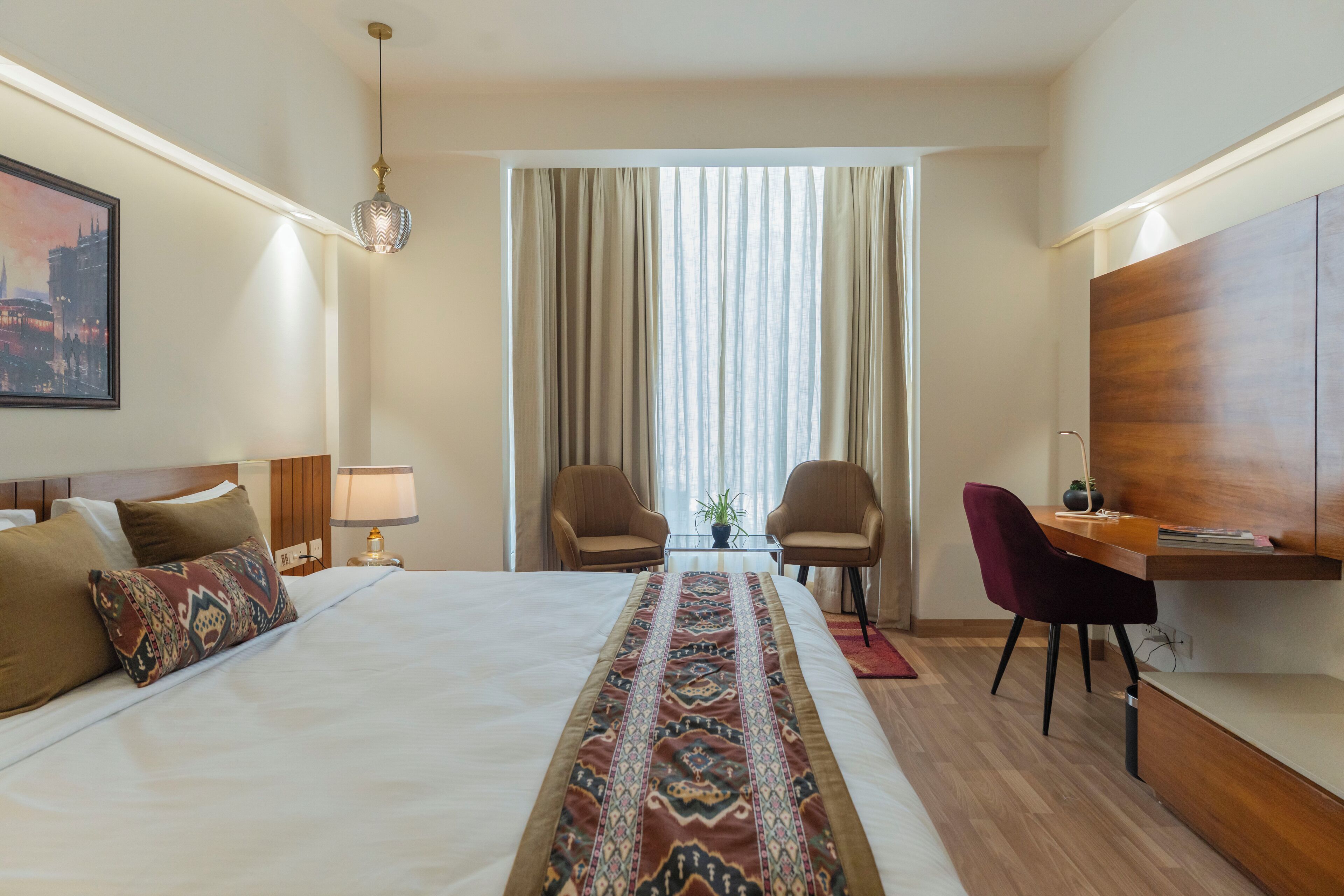A hotel room with a large bed, two chairs, a desk, and a view of the city through the window.