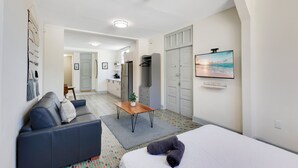 Una habitación de hotel moderna con cama, sofá, una mesita y televisor. Se observa una zona de cocina al fondo.