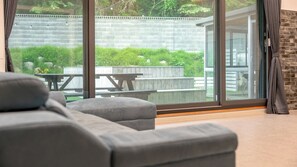 A modern living room with a grey sofa, a wooden table, and a glass door leading to an outdoor patio with a brick wall and greenery. - Gangneung Taurus Pension (Gangneung)