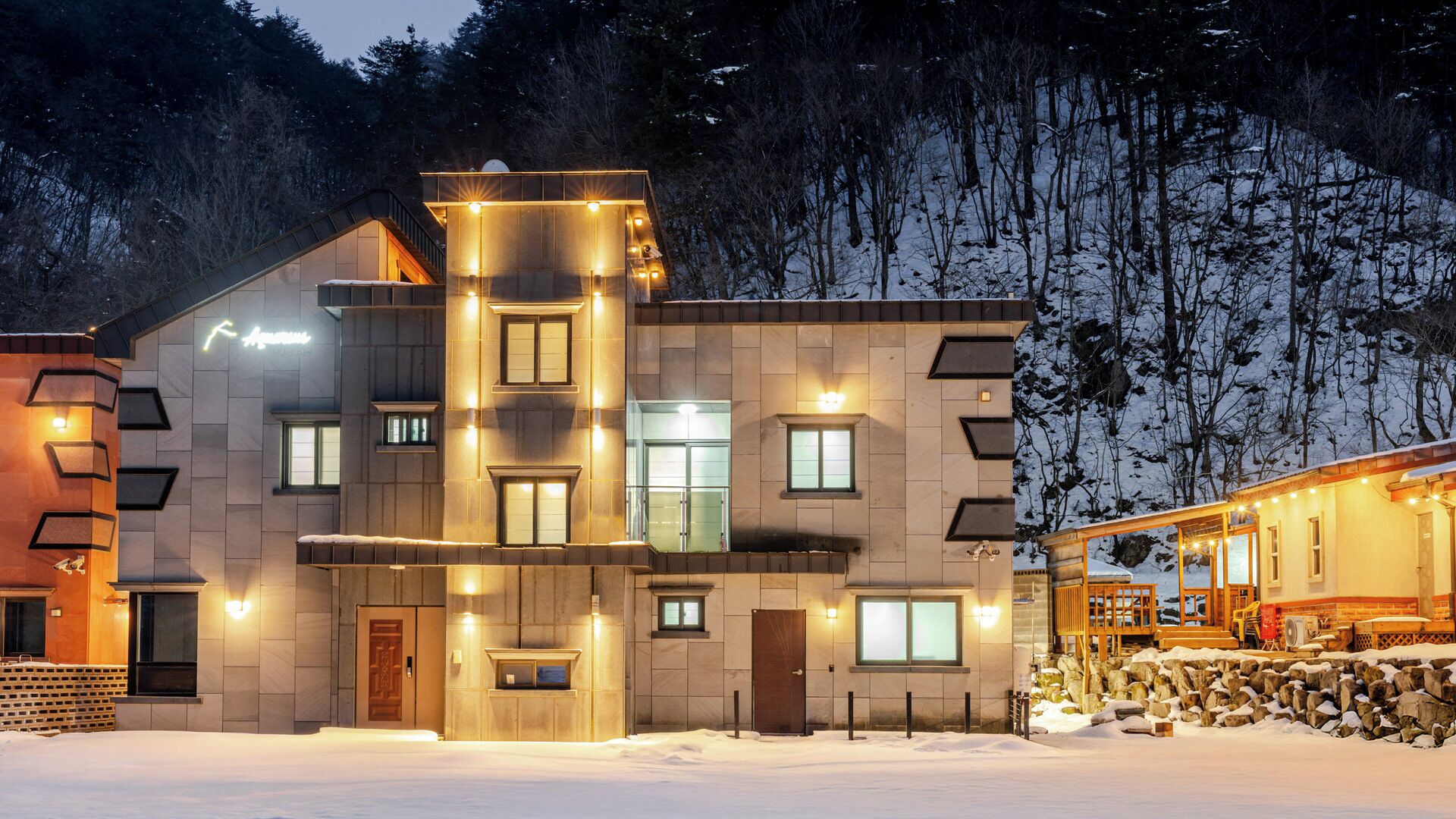 A modern building with multiple windows and a warm exterior lighting, surrounded by snow and trees.