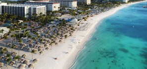 Un resort frente al mar con varios edificios hoteleros, una playa de arena bordeada de palmeras y sombrillas, y aguas turquesas cristalinas.