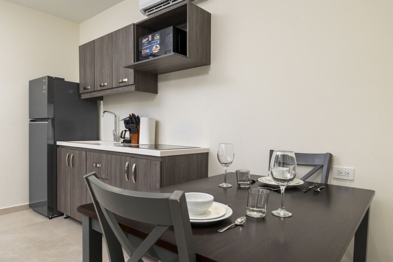 A modern kitchen with dark cabinetry, a built-in oven, and a dining table set for two with wine glasses and plates.