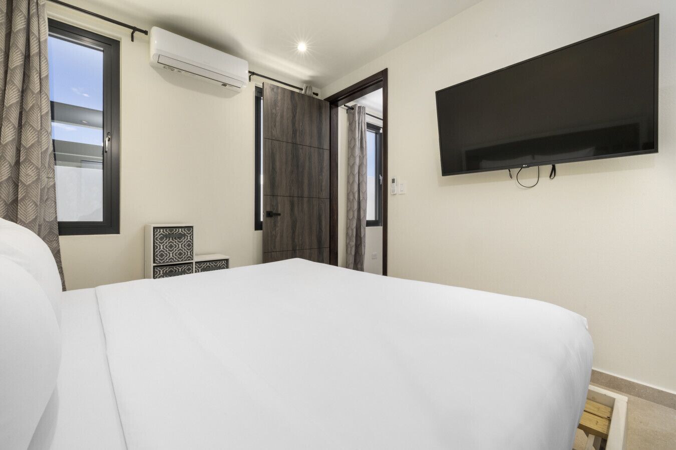 A hotel room with a large bed, a flat-screen TV mounted on the wall, a window with curtains, and an air conditioning unit.