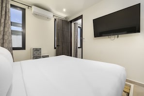A hotel room with a large bed, a flat-screen TV mounted on the wall, a window with curtains, and an air conditioning unit.