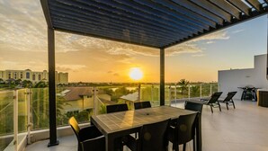 A rooftop terrace with a table and chairs, a pergola, and a view of the sunset over a cityscape.