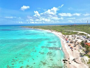 A coastal beach with clear turquoise waters, a sandy shore, and a row of thatched-roof beach huts.