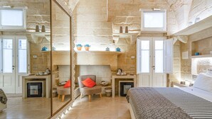 Quarto com paredes de pedra, lareira, cama com colcha listrada e espelho refletindo o ambiente.