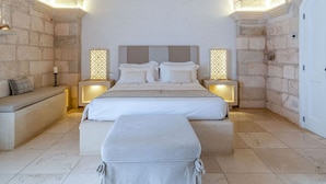 Un dormitorio con techo abovedado de piedra, una cama grande con ropa blanca, mesitas de noche con lámparas y un banco al pie de la cama.