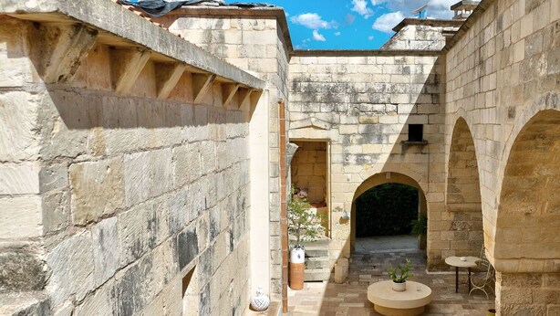 Un patio con muros de piedra, arcos y una zona central de estar con mesas y sillas.