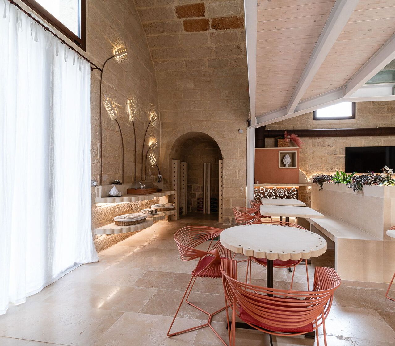 Un ambiente spazioso con parete in pietra, soffitto in legno e zona pranzo con sedie rosse e tavolo bianco.