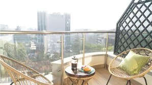 A balcony with two wicker chairs, a small round table with a coffee pot and a plate, and a view of city buildings. - Loocid Apartments at Nova (Nairobi)