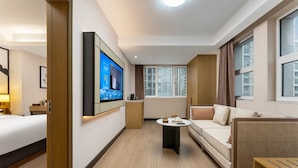 Ein Hotelzimmer mit Bett, Wandfernseher, Sofa, Couchtisch und einem Fenster mit Vorhängen.