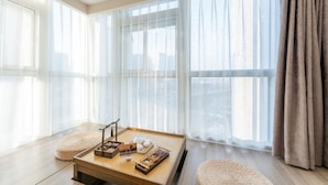 Ein Zimmer mit großen Fenstern, einem Holztisch mit Teeservice und einem gewebten Pouf.