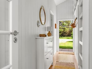 A hallway with a white door, a white dresser with drawers, a mirror, a lamp, and a view of greenery outside.