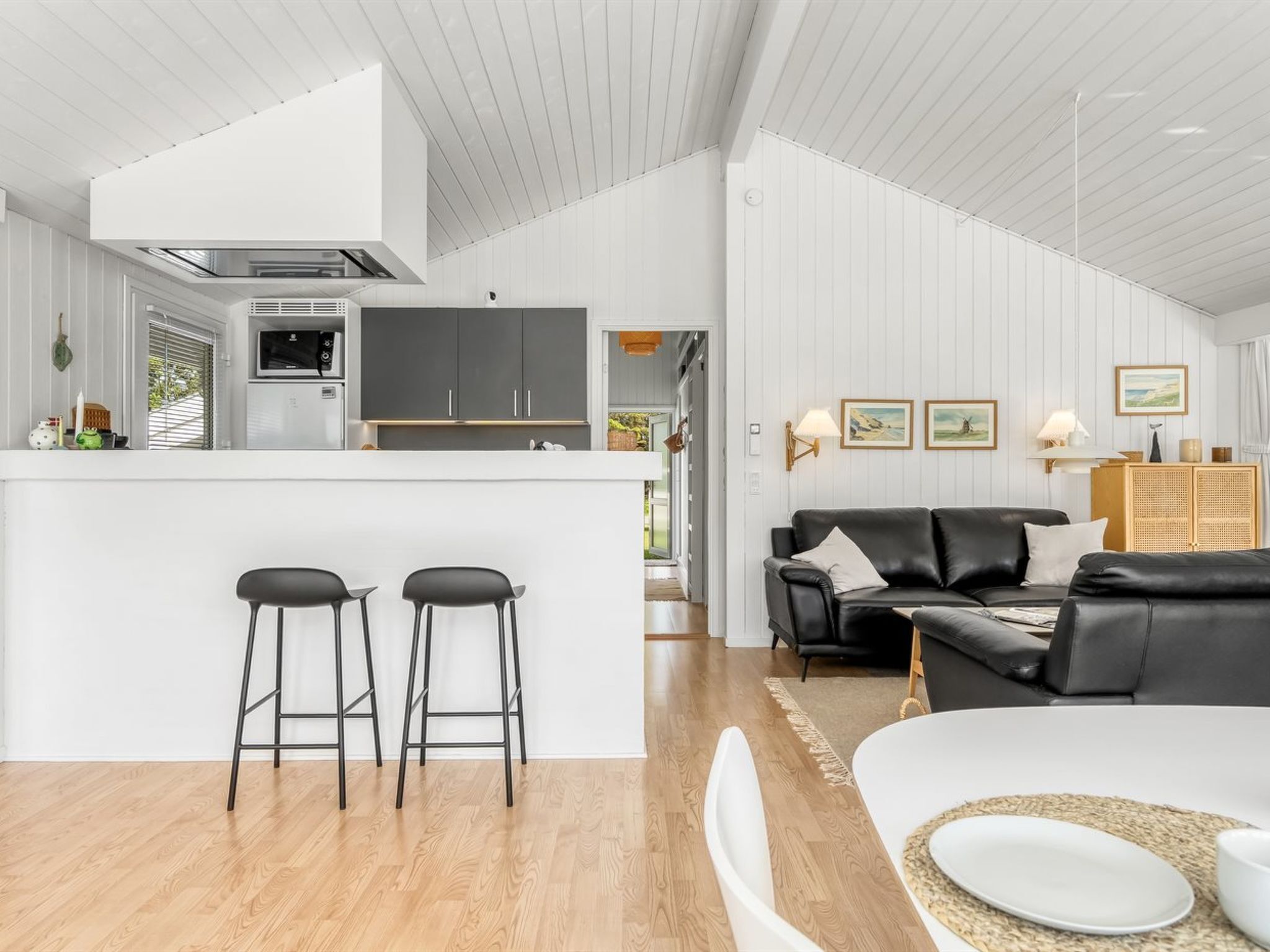 Une cuisine moderne dotée d’un îlot central, de deux tabourets de bar, d’un canapé en cuir noir et d’un coin repas avec une table et des chaises blanches.
