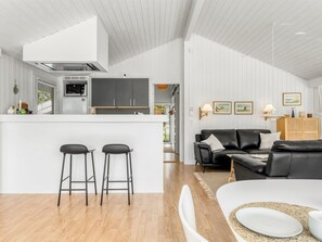 Eine moderne Küche mit Kochinsel, zwei Barhocker, einem schwarzen Ledersofa und einem Essbereich mit weißem Tisch und Stühlen.