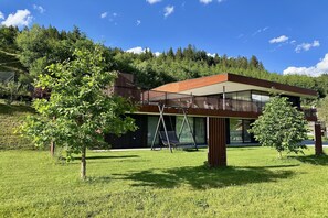 Een modern huis met een groot raam, een hangstoel en een boom in de voortuin.