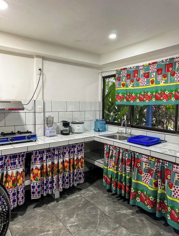 Una cocina con paredes de azulejos blancos, una estufa a gas, una licuadora, una cafetera y un lavamanos. Hay cortinas coloridas y una ventana con vista a follaje.