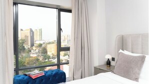 A hotel room with a large window, a bed, a bedside table with a lamp, and a blue tufted bench with books. - Loocid Apartments at Astoria (Nairobi)